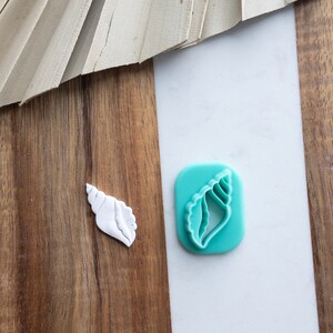 Peut inclure: Un emporte-pièce en forme de coquillage blanc et un emporte-pièce en forme de coquillage turquoise sur une surface en bois et en marbre.