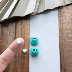 Puede incluir: Dos cortadores de galletas en forma de corazón de color turquesa sobre una superficie blanca. Un dedo con un pequeño trozo de masa en forma de corazón blanco es visible en la esquina inferior izquierda de la imagen.