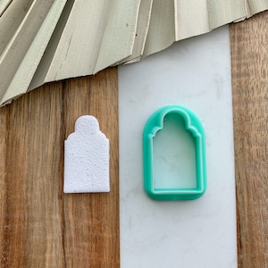 May include: A white and a green plastic cookie cutter in the shape of an arched doorway. The white cookie cutter is on a wooden surface and the green cookie cutter is on a white surface.