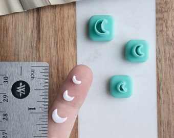 May include: Three white crescent moon shapes of varying sizes, next to a ruler, and three teal blue square-shaped molds with crescent moon cutouts.