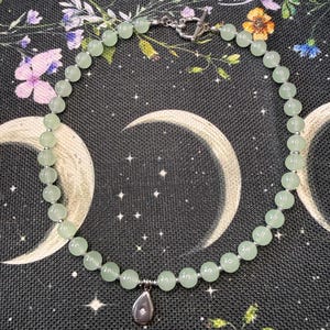 May include: A beaded necklace featuring smooth, pale green round beads interspersed with small silver-toned spacers. The necklace includes a silver-toned toggle clasp and a single silver-toned teardrop-shaped pendant with a small embedded star detail.