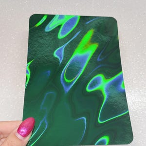 May include: A rectangular card with rounded corners, featuring a swirling pattern of green, blue, and yellow-green colors. The design has a textured appearance, with the colors blending and flowing across the surface. The card is held by a hand with pink nail polish.