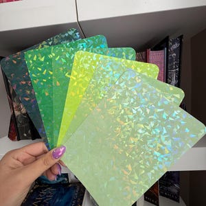 May include: A collection of rectangular cards featuring a green gradient color scheme, ranging from dark forest green to light mint green. Each card has a reflective, holographic finish with a scattered, geometric shard pattern that catches light.