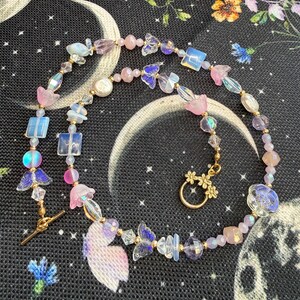 May include: A beaded necklace featuring a variety of pink, purple, and iridescent beads in shapes including butterflies, flowers, squares, and spheres. The necklace is secured with a gold-toned metal toggle clasp shaped like a small flower.