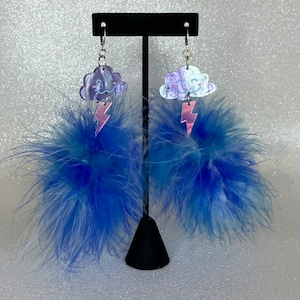 Puede incluir: Un par de pendientes caprichosos con colgantes en forma de nube con caras sonrientes y rayos rosas. Debajo de los colgantes, grandes y esponjosos adornos de plumas azules. Los pendientes cuelgan de alambres plateados.