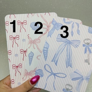 May include: Three decorative cards with bow and seashell patterns. Card 1 has pink bows and shells on a pink striped background. Card 2 has pink bows and seashells. Card 3 has blue bows and key patterns on a pink striped background.
