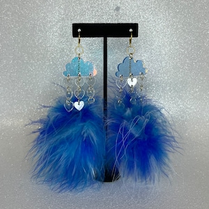 May include: A pair of blue and white feather earrings with a cloud and heart design. The earrings are made of iridescent plastic and have a silver finish.