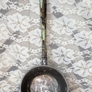 May include: Vintage metal cooking pot with a long wooden handle. The handle has a distressed, aged appearance with shades of green and brown. The pot itself is a dull silver color, showing signs of wear.