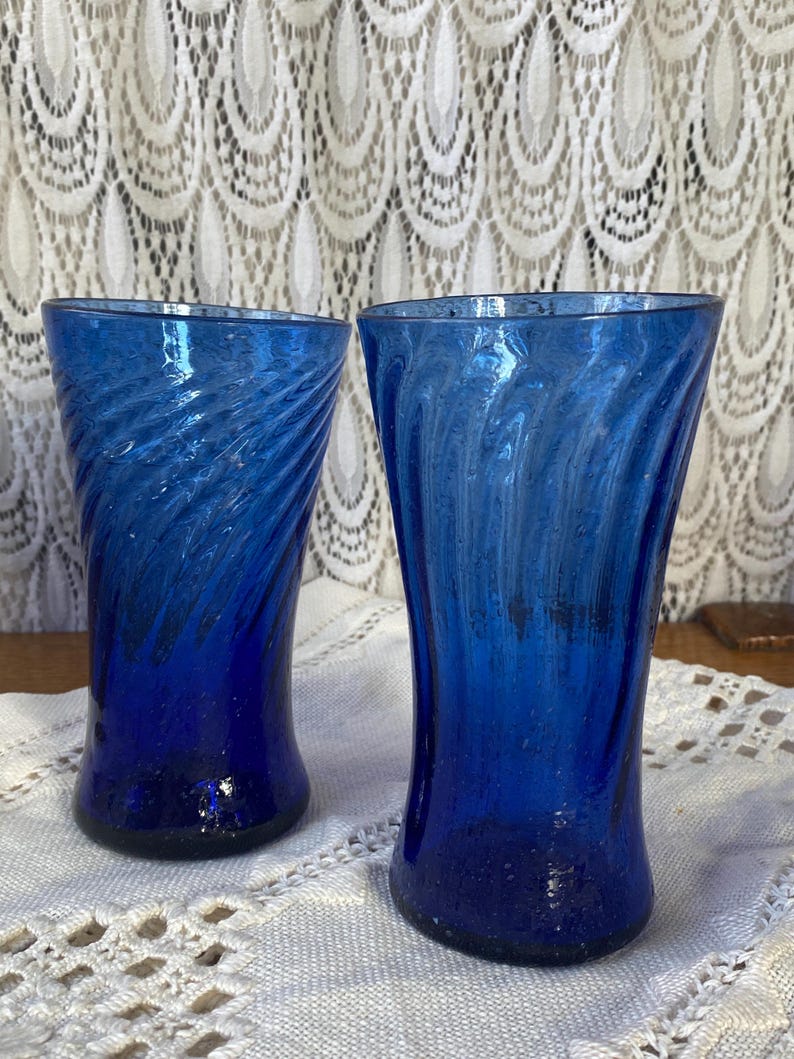 Pu&ograve; includere: Due bicchieri di vetro blu cobalto con un design a spirale. I bicchieri hanno una forma svasata e poggiano su una tovaglia di pizzo bianco. Lo sfondo presenta una tenda di pizzo bianco.