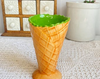 Può includere: Un vaso in ceramica a forma di cono gelato con esterno arancione strutturato e interno verde brillante. Il vaso ha un bordo smerlato e una base rotonda. Lo sfondo include una rastrelliera per spezie in legno e un vaso in ceramica bianca.