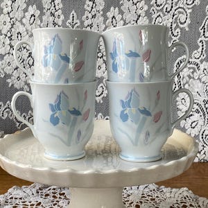 Może przedstawiać: Cztery białe ceramiczne kubki do kawy ułożone na białym stojaku na ciasto. Każdy kubek ma kwiatowy wzór z niebieskimi, różowymi i zielonymi akcentami. Kubki mają zakrzywiony uchwyt i lekko rozszerzoną krawędź. Stojak na ciasto ma falisty brzeg.