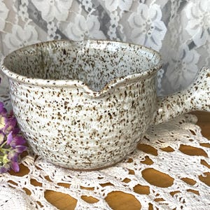 May include: A speckled ceramic bowl with a handle. The bowl is off-white with brown speckles and a pouring spout. The handle is thick and textured. The bowl sits on a white lace doily, with purple flowers in the background.