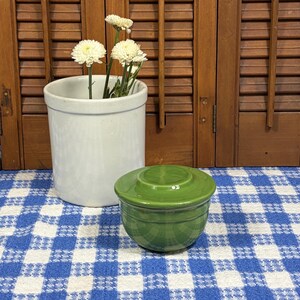 Peut inclure: Un petit pot &agrave; beurre en c&eacute;ramique vert brillant avec un couvercle assorti, plac&eacute; &agrave; c&ocirc;t&eacute; d'un vase cylindrique en c&eacute;ramique blanche contenant des chrysanth&egrave;mes blancs. Les objets sont pos&eacute;s sur un tissu &agrave; carreaux bleus et blancs.