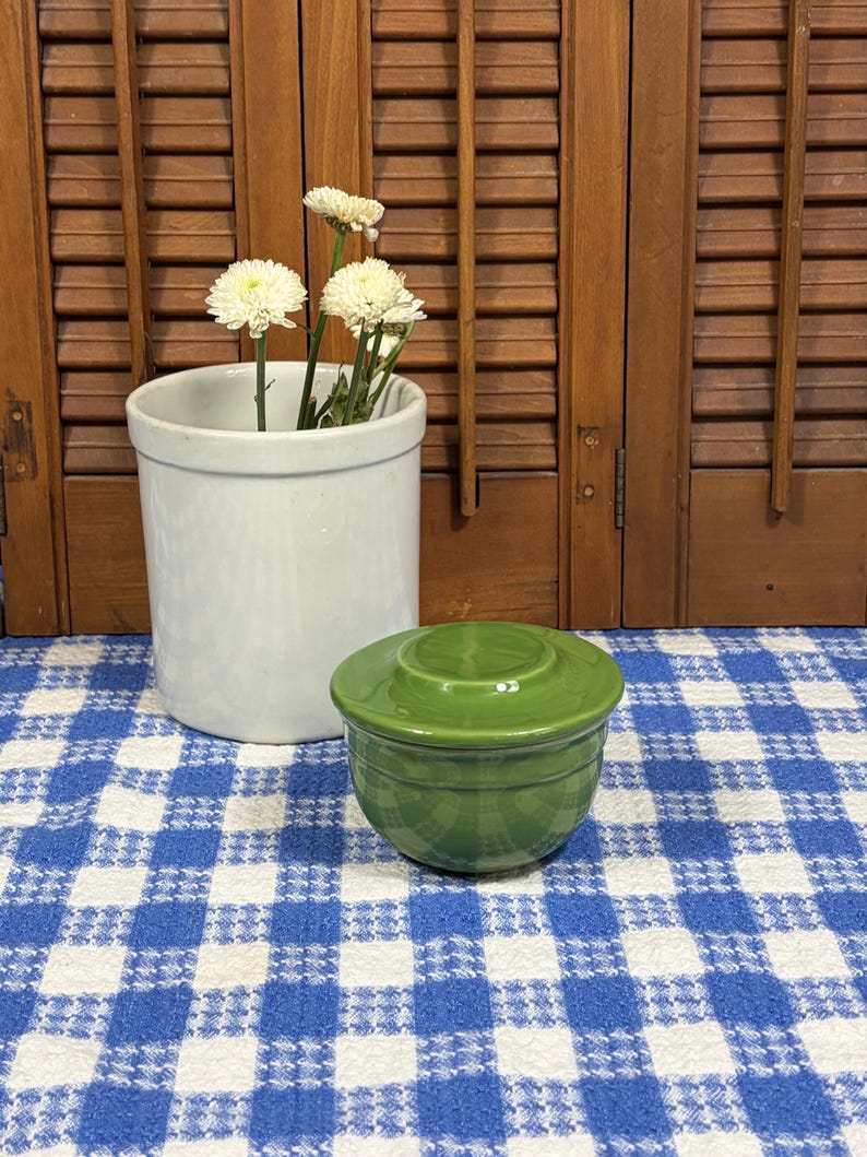 Peut inclure: Un petit pot &agrave; beurre en c&eacute;ramique vert brillant avec un couvercle assorti, plac&eacute; &agrave; c&ocirc;t&eacute; d'un vase cylindrique en c&eacute;ramique blanche contenant des chrysanth&egrave;mes blancs. Les objets sont pos&eacute;s sur un tissu &agrave; carreaux bleus et blancs.