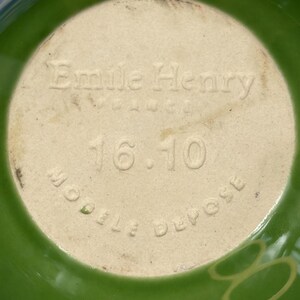 Peut inclure: Le dessous d'un bol en c&eacute;ramique verte pr&eacute;sentant le texte en relief Emile Henry, France, 16.10 et Modele Depose. La base est de couleur beige clair, contrastant avec l'&eacute;mail vert vif qui recouvre l'ext&eacute;rieur du bol.