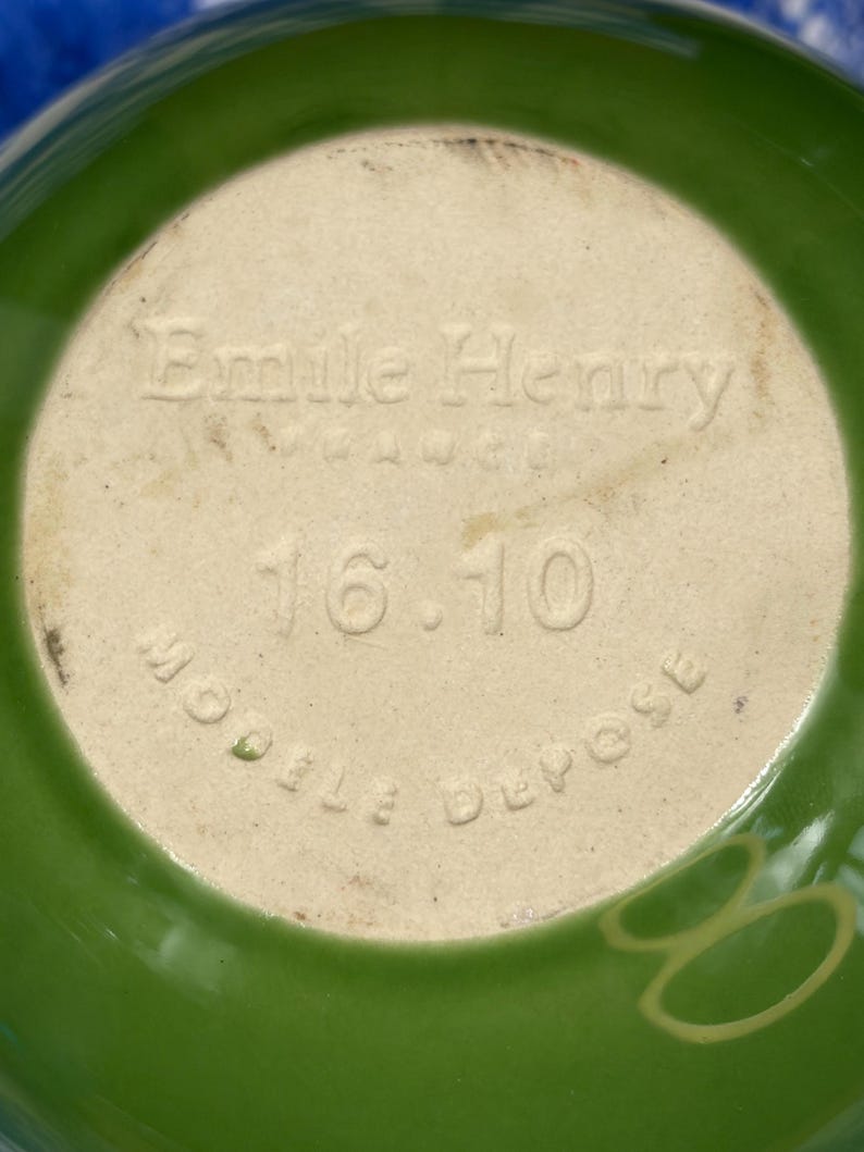 Peut inclure: Le dessous d'un bol en c&eacute;ramique verte pr&eacute;sentant le texte en relief Emile Henry, France, 16.10 et Modele Depose. La base est de couleur beige clair, contrastant avec l'&eacute;mail vert vif qui recouvre l'ext&eacute;rieur du bol.