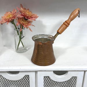 May include: A copper coffee pot with a hammered texture, a wide base, and a tapered body. It features a long, turned wooden handle attached by a brass-colored metal joint and a small pouring spout at the rim. The interior is lined with silver-colored metal.