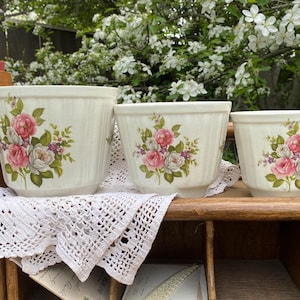 Puede incluir: Conjunto de tres macetas de cerámica blanquecina con textura acanalada. Cada maceta presenta un diseño floral con rosas rosas y blancas, detalles morados y hojas verdes. Las macetas están dispuestas sobre una superficie de madera con un tapete de encaje blanco.