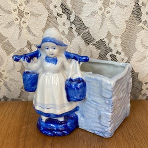 May include: A blue and white ceramic planter featuring a Dutch girl figurine carrying buckets. The planter has a rectangular shape with a textured, wood-like appearance. The figurine is in a traditional Dutch outfit.
