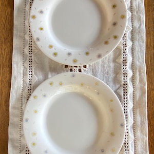May include: Two white ceramic plates with a delicate floral pattern of yellow and blue flowers. The plates are arranged on a white lace runner, creating a charming and elegant aesthetic. The plates are perfect for serving meals.