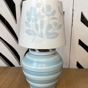 May include: A small, decorative lamp with a light blue and white color scheme. The base is a rounded, striped ceramic design. The lampshade is white with a floral pattern in light blue. The lamp is on a wooden surface.