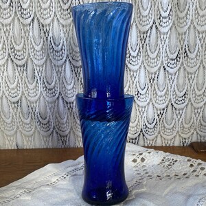 Pu&ograve; includere: Due vasi di vetro blu cobalto con un design a spirale. I vasi sono impilati, con il vaso superiore leggermente pi&ugrave; alto di quello inferiore. Lo sfondo presenta una tenda di pizzo bianco.
