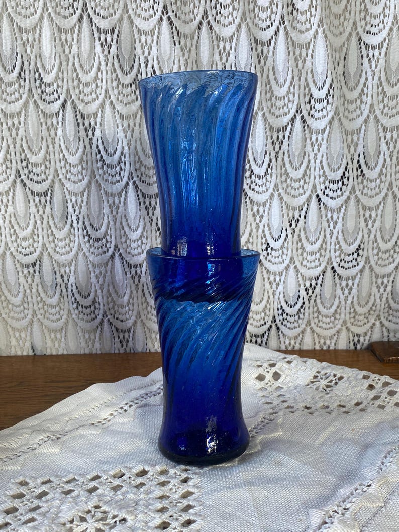 Pu&ograve; includere: Due vasi di vetro blu cobalto con un design a spirale. I vasi sono impilati, con il vaso superiore leggermente pi&ugrave; alto di quello inferiore. Lo sfondo presenta una tenda di pizzo bianco.