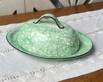 Può includere: Una burriera in ceramica con un motivo maculato verde e bianco, con coperchio e manico nero. Il piatto è posto su un panno bianco con bordi in pizzo, su una superficie in legno.