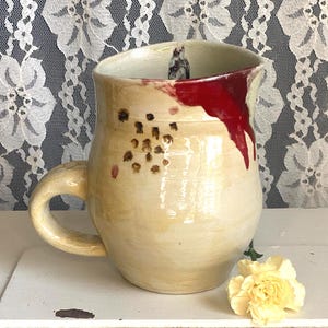 Könnte beinhalten: Handgefertigte Keramik-Tasse mit cremefarbenem Boden, großem Henkel und roter Glasur, die an der Seite herunterläuft. Die Tasse hat braune und rote gesprenkelte Details und eine kleine Figur im Inneren. Eine gelbe Blume liegt in der Nähe der Tasse.