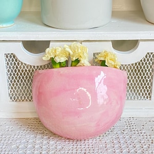 Puede incluir: Un jarrón de pared de cerámica rosa con efecto marmolado, que contiene varias flores amarillas. El jarrón tiene forma redondeada y se muestra sobre un fondo blanco. Las flores añaden un toque de color al jarrón rosa suave.