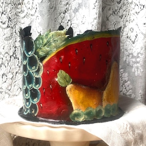 May include: A hand-painted ceramic container with a watermelon design. The artwork features red, green, and black colours, with a black base. The container is displayed on a white cake stand.