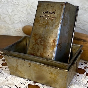 Puede incluir: Dos moldes de pan de metal vintage, uno dentro del otro. Los moldes tienen un aspecto desgastado, con óxido y decoloración visibles. El molde superior tiene el texto en relieve "prit ated" y "AZUR IN ADAM". Sobre un mantel de encaje blanco.