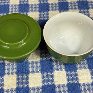 Peut inclure: Un ensemble de beurrier en c&eacute;ramique compos&eacute; de deux pi&egrave;ces avec un ext&eacute;rieur vert olive uni. Une pi&egrave;ce est un couvercle circulaire avec une poign&eacute;e centrale sur&eacute;lev&eacute;e, et l'autre est un bol ouvert et vide avec un int&eacute;rieur blanc brillant et lisse.