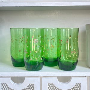 Puede incluir: Cuatro vasos verdes con un diseño floral. Los vasos tienen una base verde oscuro y están decorados con pequeñas flores de colores. Los vasos se exhiben en una estantería blanca.