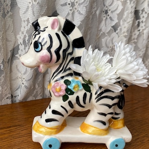 Könnte beinhalten: Ein Keramik-Zebra-Pflanzgefäß mit schwarz-weißen Streifen, rosa Wangen und blauen Augen. Das Zebra ist mit bunten Blumen verziert und steht auf einem weißen Sockel mit blauen Rädern. Weiße Blumen sind im Rücken des Zebras platziert.