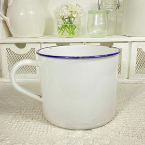 May include: A white enamel mug with a single handle and a thin blue rim. The mug features a smooth, glossy finish with visible signs of wear along the top edge, showing a metallic base underneath the white coating on the rim of the cup.