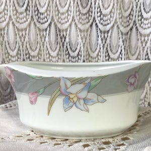May include: A white porcelain serving bowl with a gray band featuring a floral design of pink and blue flowers. The bowl has a curved shape and is set against a lace background.