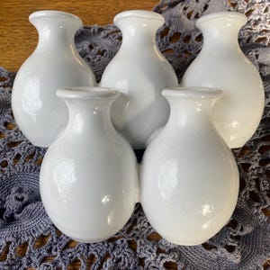 Może przedstawiać: Pięć małych, białych wazonów ceramicznych o zaokrąglonym korpusie i wąskiej szyi. Wazony są ułożone blisko siebie na szarej koronkowej serwetce, tworząc prostą, elegancką ekspozycję. Wszystkie wazony mają ten sam rozmiar i kształt.