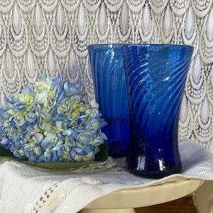 Pu&ograve; includere: Due bicchieri di vetro blu cobalto con un design a spirale. Un bouquet di ortensie blu e gialle poggia su un panno bianco. I bicchieri sono su un piedistallo bianco, con una tenda di pizzo bianco sullo sfondo.