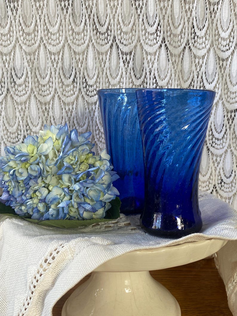 Pu&ograve; includere: Due bicchieri di vetro blu cobalto con un design a spirale. Un bouquet di ortensie blu e gialle poggia su un panno bianco. I bicchieri sono su un piedistallo bianco, con una tenda di pizzo bianco sullo sfondo.