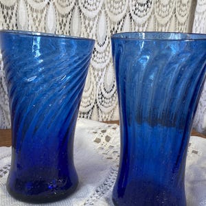 Pu&ograve; includere: Due bicchieri blu cobalto con un design a spirale. I bicchieri sono in vetro e hanno una superficie strutturata. I bicchieri sono alti e cilindrici. I bicchieri sono su una tovaglia di pizzo bianco. Ogni bicchiere &egrave; alto circa 15 cm.