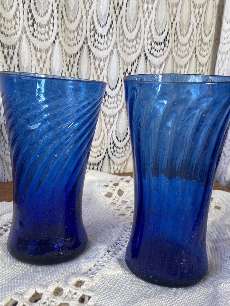 Pu&ograve; includere: Due bicchieri blu cobalto con un design a spirale. I bicchieri sono in vetro e hanno una superficie strutturata. I bicchieri sono alti e cilindrici. I bicchieri sono su una tovaglia di pizzo bianco. Ogni bicchiere &egrave; alto circa 15 cm.