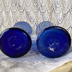 Pu&ograve; includere: Due vasi di vetro blu cobalto con base rotonda e design a righe. I vasi sono appoggiati su uno sfondo di pizzo bianco. I fondi dei vasi sono a fuoco, mostrando il colore blu intenso.
