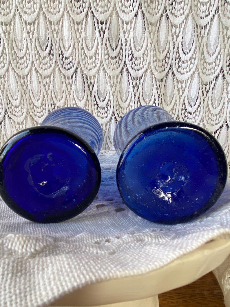 Pu&ograve; includere: Due vasi di vetro blu cobalto con base rotonda e design a righe. I vasi sono appoggiati su uno sfondo di pizzo bianco. I fondi dei vasi sono a fuoco, mostrando il colore blu intenso.