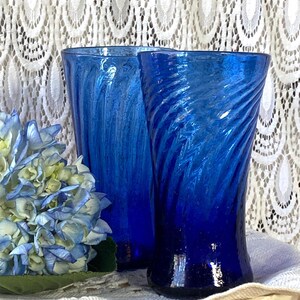 Pu&ograve; includere: Due vasi di vetro blu cobalto con un design a spirale. I vasi sono accanto a un bouquet di ortensie blu e bianche. Lo sfondo &egrave; una tenda di pizzo bianco.
