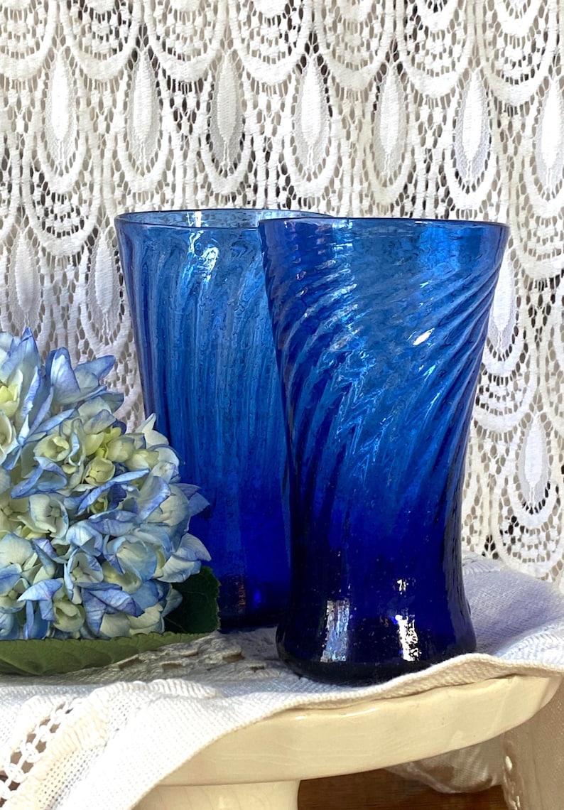 Pu&ograve; includere: Due vasi di vetro blu cobalto con un design a spirale. I vasi sono accanto a un bouquet di ortensie blu e bianche. Lo sfondo &egrave; una tenda di pizzo bianco.