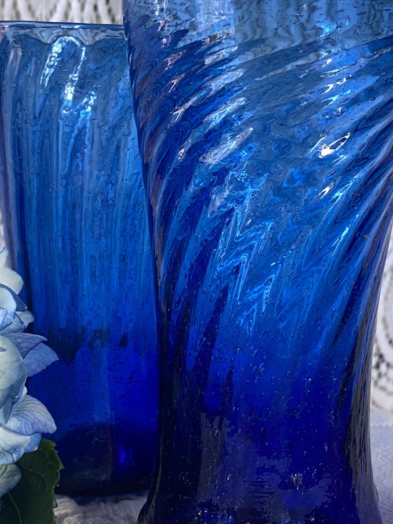 Pu&ograve; includere: Due alti bicchieri di vetro blu cobalto con un design testurizzato e a spirale. I bicchieri hanno un'estetica vintage e sono parzialmente visibili, mostrando il loro motivo decorativo unico. La tonalit&agrave; blu &egrave; ricca e vibrante.