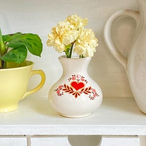Könnte beinhalten: Eine kleine, weiße Keramikvase mit einem roten Herz- und Blumendesign, die mehrere gelbe Blumen enthält. Die Vase steht auf einem weißen Regal, mit einer gelben Tasse und einer weißen Kanne im Hintergrund.