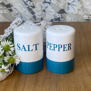 May include: A pair of ceramic salt and pepper shakers. The shakers are white with a teal blue base and the words "SALT" and "PEPPER" in teal blue lettering. Each shaker has three holes on top.