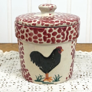May include: A cream-colored ceramic canister with a matching lid, featuring a hand-painted black rooster standing on green grass. The rim of the lid and the upper section of the canister are decorated with a pattern of small, irregular red spots.